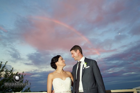 A sun shower at sunset makes for a beautiful rainbow, and the bride and groom take a break from greeting guests for a portrait on the balcony, reception at Geneva National Golf Club. Wedding photography by Steve & Tiffany Warmowski